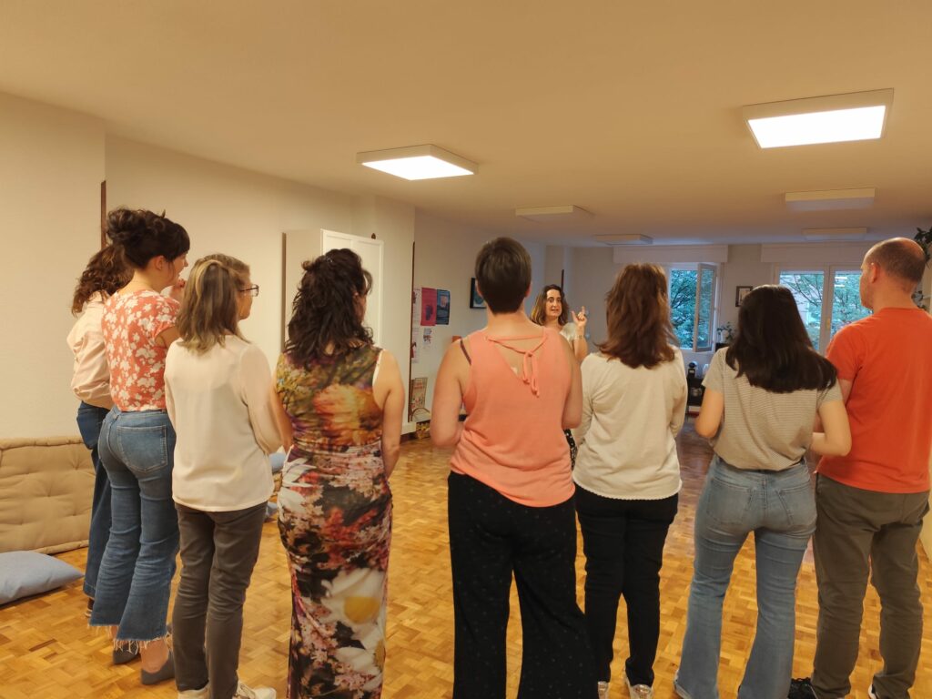 Grupo de personas de pie en corro escuchando a una mujer que habla en una sala luminosa con suelo de parquet