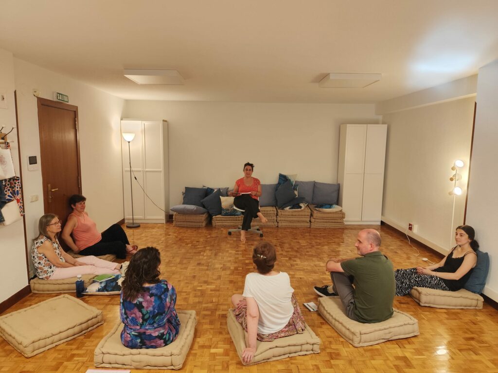 Sesión de grupo Gestalt: una mujer en una silla habla a seis personas sentadas en cojines sobre parquet