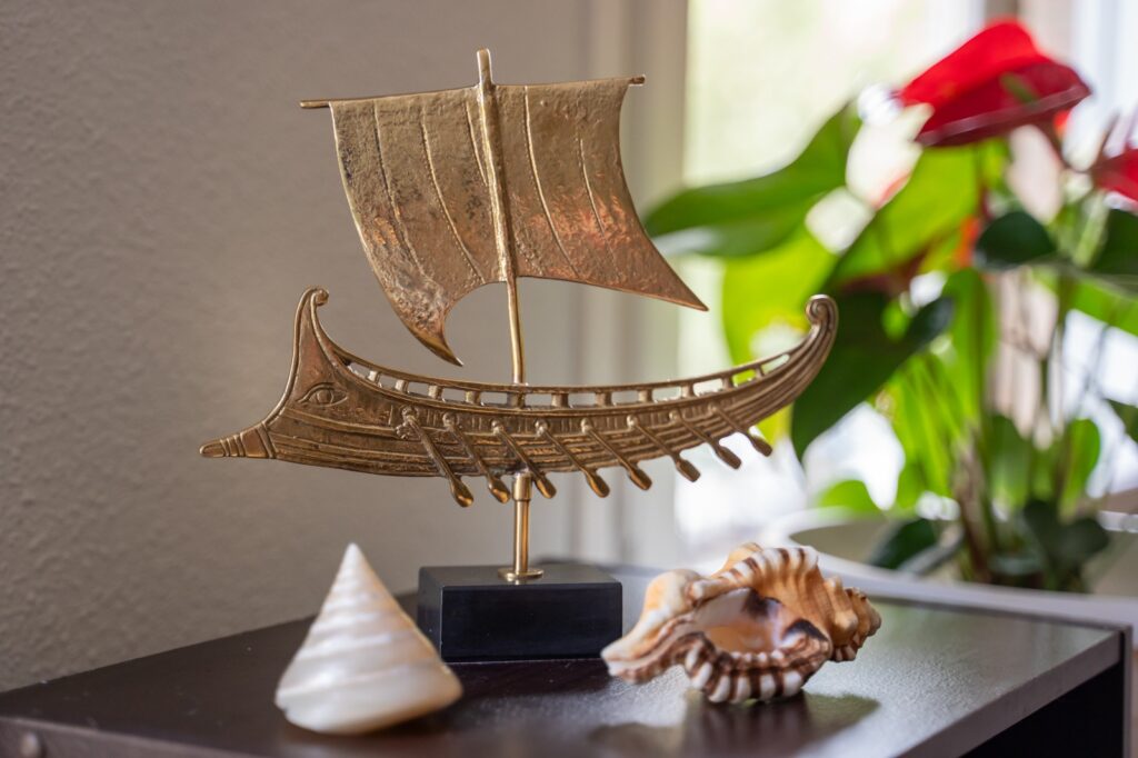 Figura de latón de un barco griego antiguo con vela y remos sobre una peana negra, junto a dos caracolas marinas